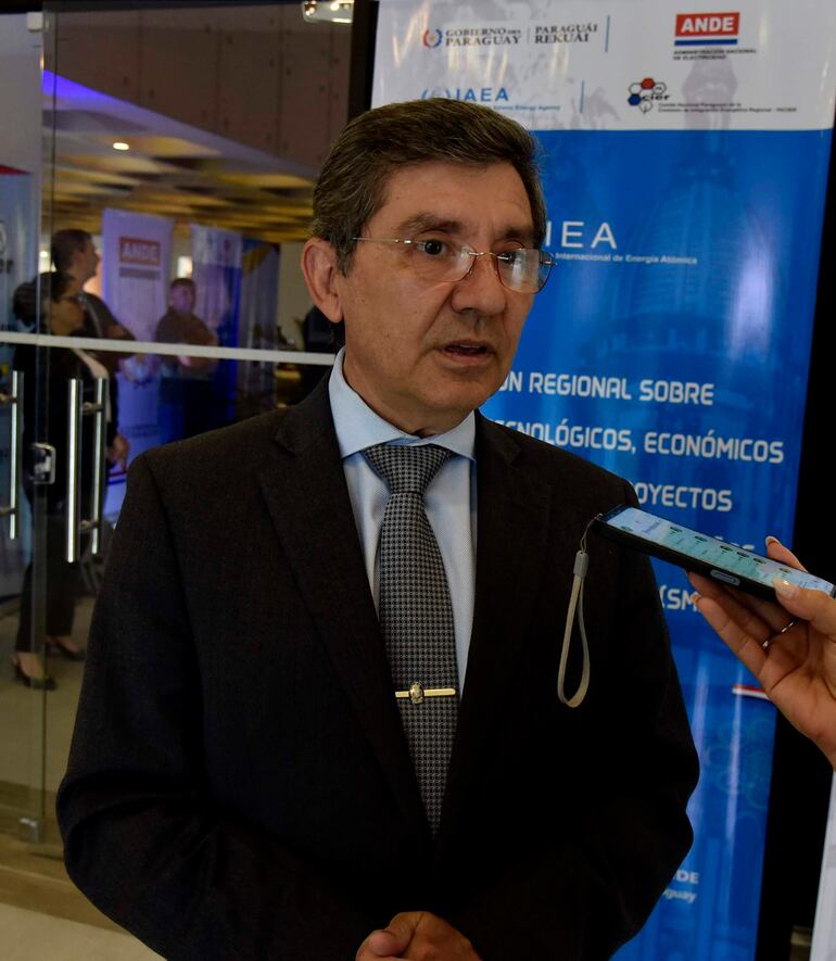 Hombre de cabello corto y canoso con traje negro y corbata, entrevistado en evento formal con asistentes al fondo.