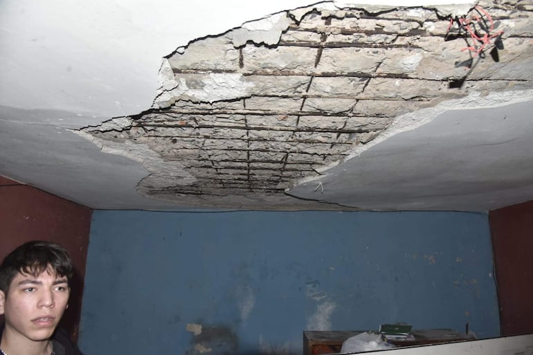 Joven preocupado inspecciona un techo deteriorado con fisuras visibles en una habitación descuidada.