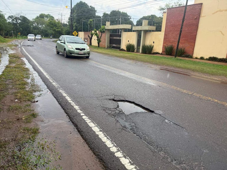 Tres vehículos, incluyendo un auto verde claro, pasan por un bache con agua acumulada en la carretera, rodeados de vegetación.