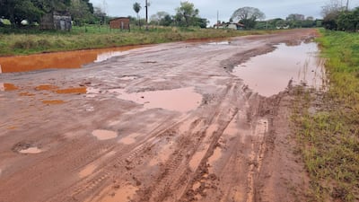 Carretera de tierra con surcos y charcos de agua, rodeada de vegetación y casas simples en un día nublado.