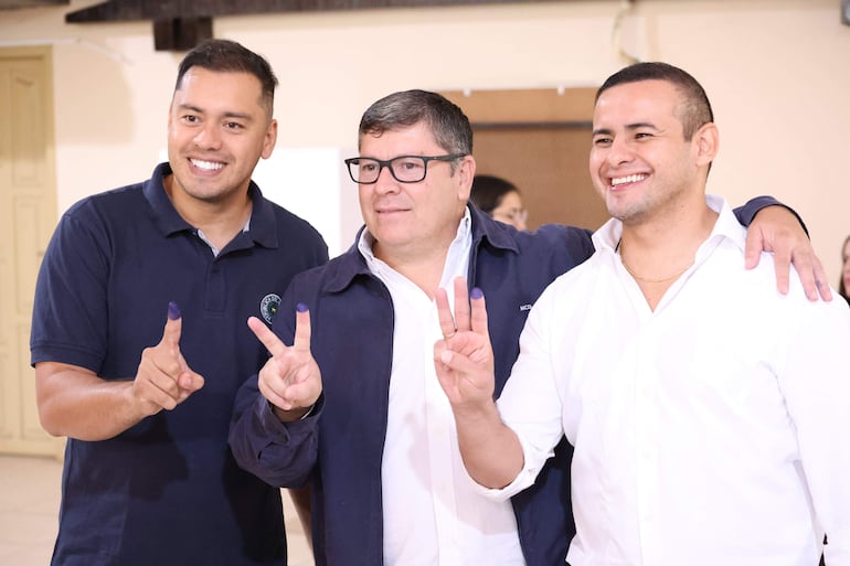 Daniel Pereira Mujica y dos hombres sonrientes en interior, con chaqueta oscura, gestos de victoria y dedos manchados de tinta.