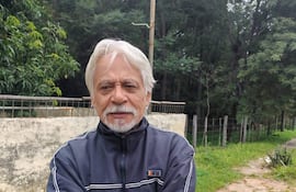 Hombre mayor con chaqueta azul oscura y barba blanca, cruzando los brazos en un ambiente natural con vegetación.