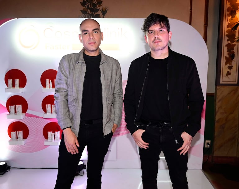 Dos hombres con chaqueta gris y abrigo negro posan juntos ante estantes de productos CosmeClinik en un ambiente iluminado.