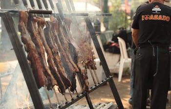 Hombre de espaldas con camiseta 'I ❤️ ASADO' asando carne al aire libre rodeado de humo y ambiente festivo.