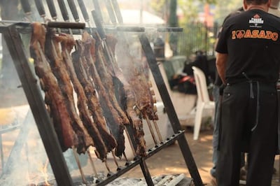 Hombre de espaldas con camiseta 'I ❤️ ASADO' asando carne al aire libre rodeado de humo y ambiente festivo.