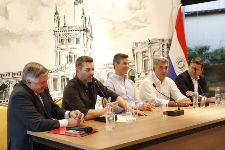 Santiago Peña Santi sonríe mientras con un micrófono, otros hombres escuchan atentamente en un ambiente de conferencia formal.
