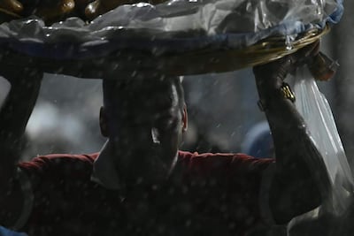 Hombre con camiseta roja y cuello blanco sostiene una bandeja sobre su cabeza en un ambiente lluvioso.