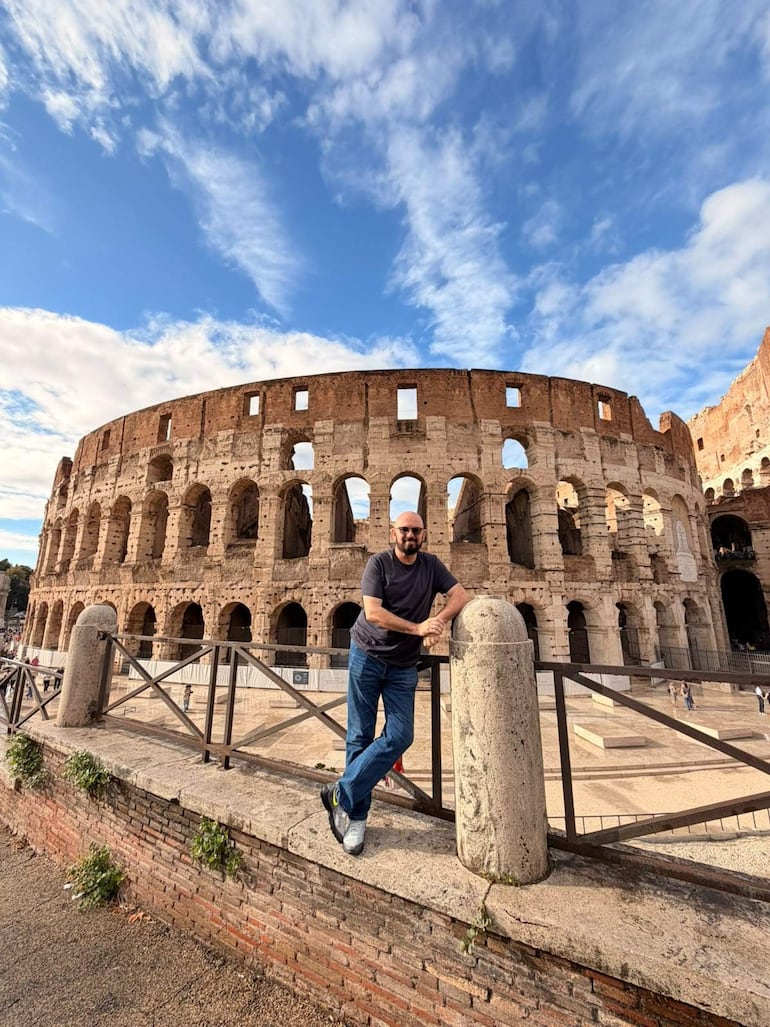 Hombre con cabello oscuro y barba, viste camiseta gris y jeans, frente al Coliseo con cielo nublado.