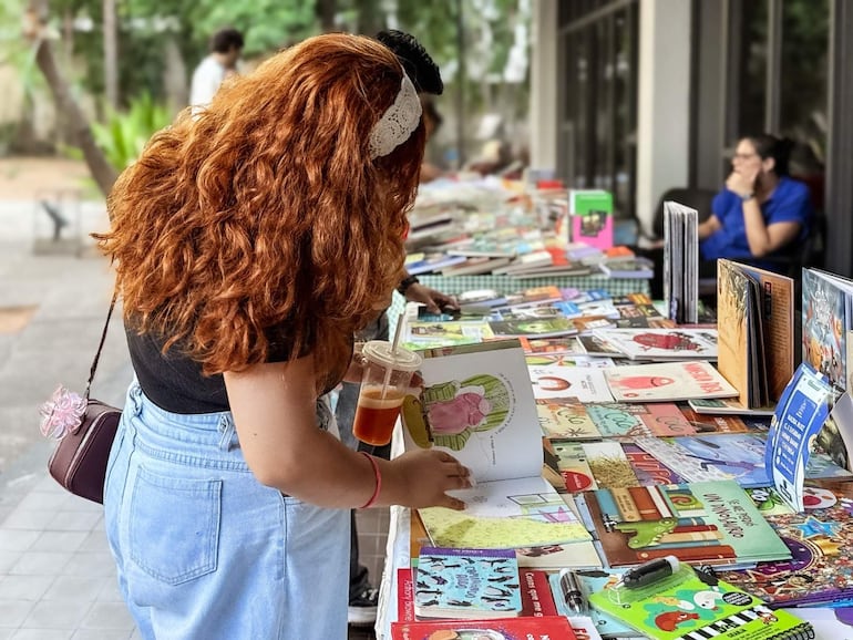 Mujer de cabello rizado y rojizo en camiseta negra revisa libros en un puesto, sosteniendo un vaso de bebida en un ambiente natural.