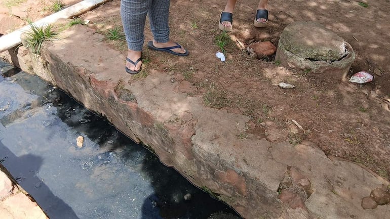 Dos personas, una con pantalones de cuadro y sandalias, y otra en camiseta y sandalias, observan el agua oscura de un desagüe.