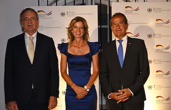 Mujer sonriente con vestido azul y dos hombres con trajes oscuros, posando en un ambiente formal con logotipos de la embajada.