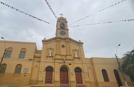Iglesia amarilla con torre central y reloj, flanqueada por estatuas, bajo un cielo nublado y banderas festivas.