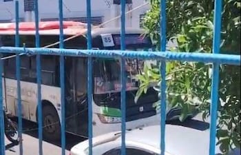 Autobús blanco con detalles verdes de la línea 85 y automóvil claro estacionado, observados desde detrás de una reja azul.