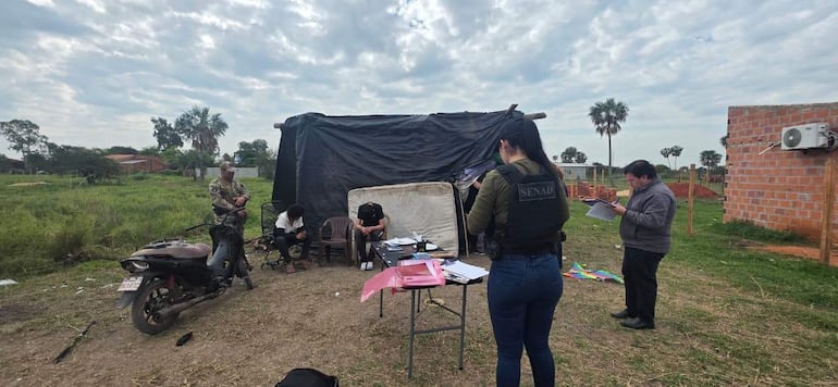 Mujer de espaldas con chaleco 'SENAD', hombres sentados y de pie con papeles en un refugio de plástico negro.