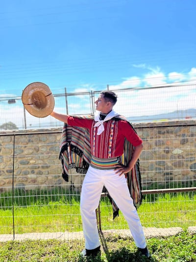 Brahian Barrios, vestido con camisa roja y poncho colorido, sostiene un sombrero mientras posa en un campo.