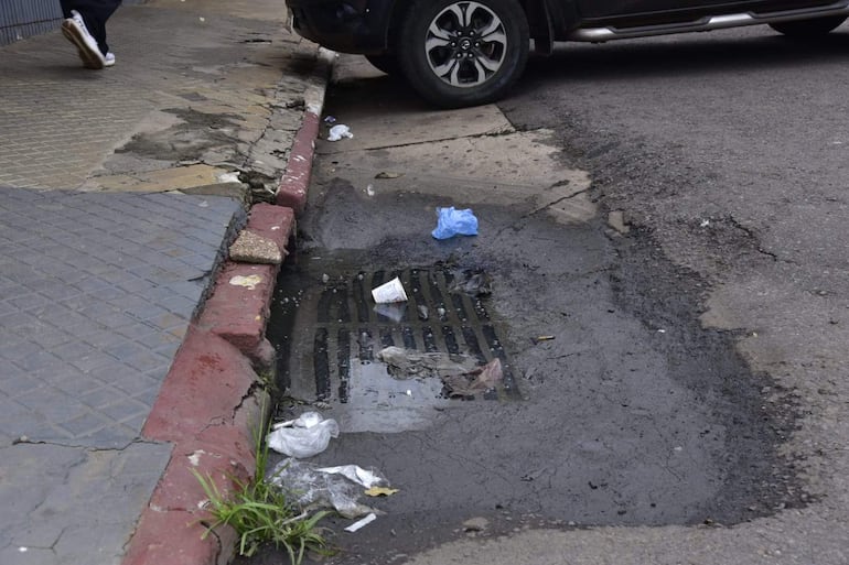 Una calle de Asunción muestra un bache lleno de agua y basura, con un vehículo estacionado y vereda en mal estado a los lados.