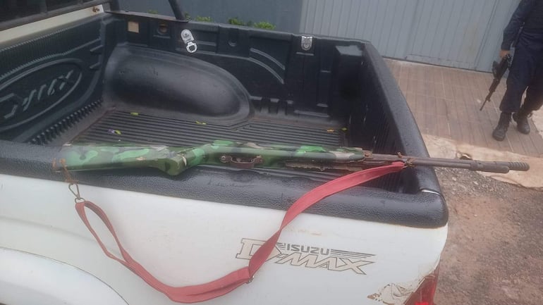 Rifle camuflado apoyado en la caja de una camioneta blanca, con un oficial armado parcialmente visible en uniforme oscuro.