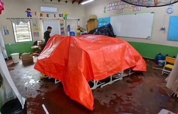 Persona de pie en aula desordenada, vestimenta oscura, observando mesas bajo un toldo rojo. Paredes con decoraciones educativas y piso húmedo.