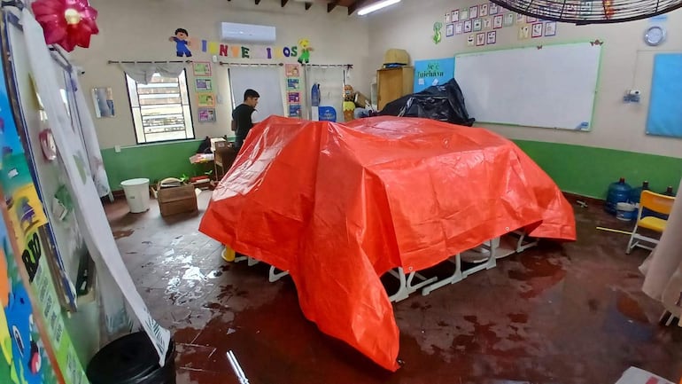 Persona de pie en aula desordenada, vestimenta oscura, observando mesas bajo un toldo rojo. Paredes con decoraciones educativas y piso húmedo.