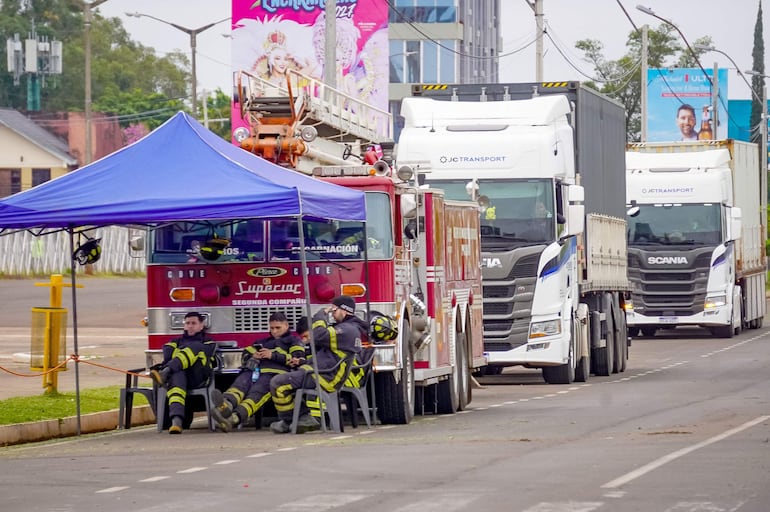 Tres bomberos en uniformes grises con detalles amarillos se relajan sentados frente a una camioneta roja bajo una carpa azul.