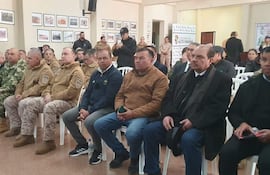 Hombres en atuendo formal y militar conversan en evento oficial, con ambiente tranquilo y decoraciones en las paredes.