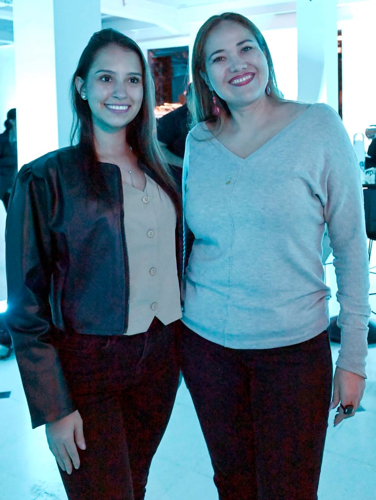 Mujer con chaqueta negra y blusa clara, junto a otra con suéter gris, sonriendo en evento iluminado con luces azuladas.