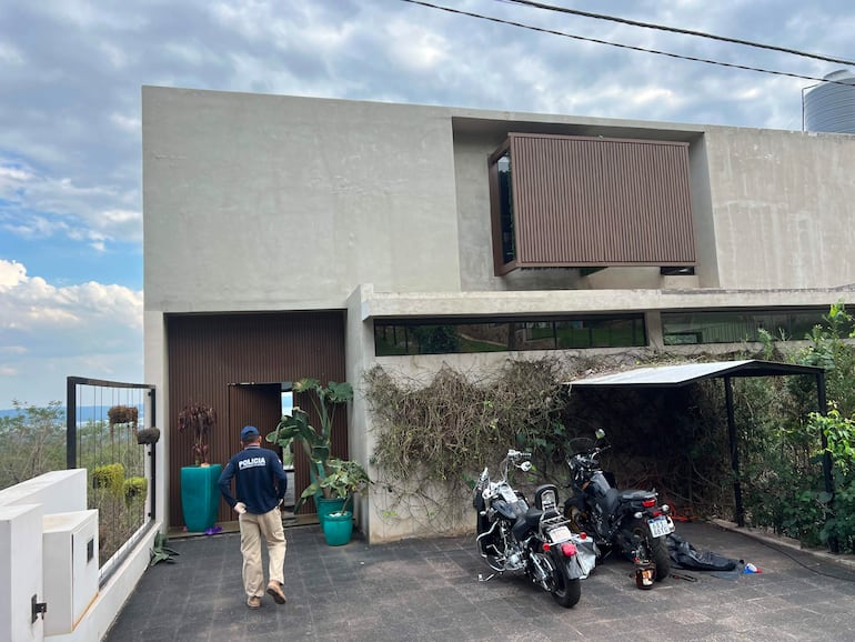 Policía con uniforme camina hacia la entrada de una vivienda moderna, con motocicletas y plantas visibles en el entorno.