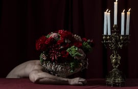 Mesa con mantel rojo, candelabro encendido y mano junto a un jarrón con flores rojas y verdes, en ambiente sombrío.