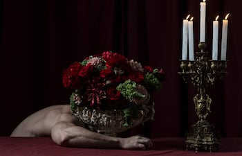 Mesa con mantel rojo, candelabro encendido y mano junto a un jarrón con flores rojas y verdes, en ambiente sombrío.