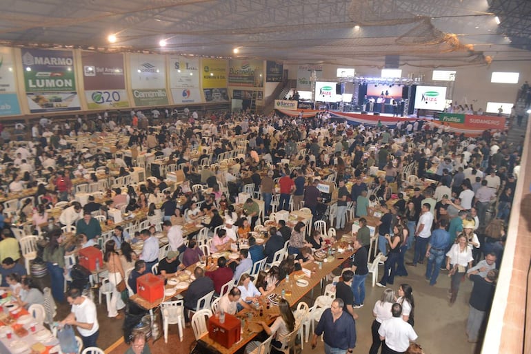 Multitud en formal, sentados en mesas, disfrutando de comida y bebida en un ambiente festivo, con un escenario al fondo.