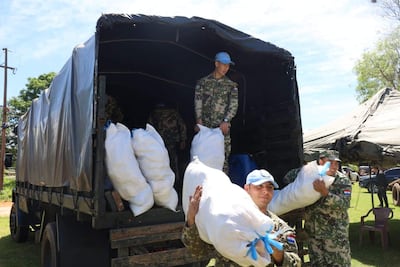 Cinco soldados en uniformes de camuflaje cargan costales cerca de un camión en un área boscosa y de césped.