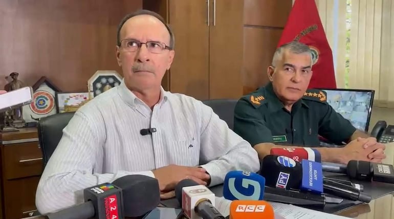 Óscar González en camisa blanca y gafas, junto a un oficial militar en uniforme verde, ambos en un ambiente de oficina con micrófonos.