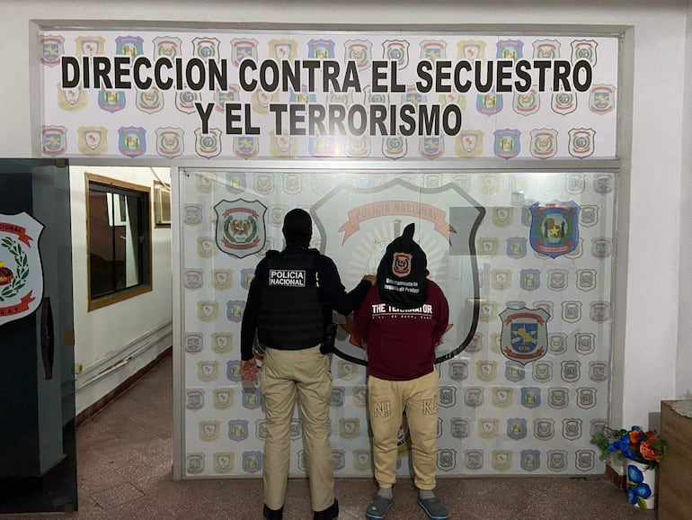 Hombre arrestado con camiseta roja 'THE TERMINATOR' frente a un policía en un ambiente institucional con escudo de la policía.