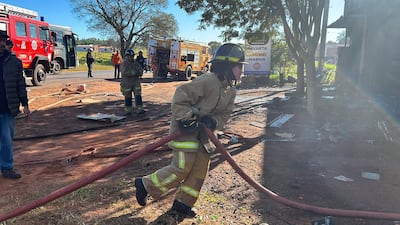 Bombero en acción con manguera, equipado con casco y traje, mientras otros bomberos lo asisten en un entorno lleno de escombros.