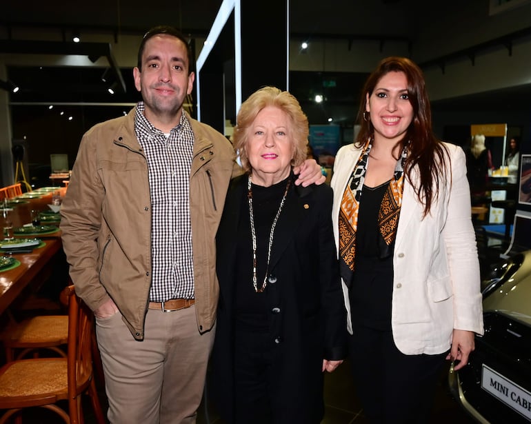Tres personas sonríen en un elegante evento: hombre en chaqueta beige, mujer rubia en atuendo negro y mujer en blanco con bufanda colorida.