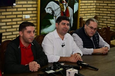 Magno Álvarez con camiseta roja y chaqueta negra en mesa con micrófonos, rodeado de dos hombres con vestimenta formal.