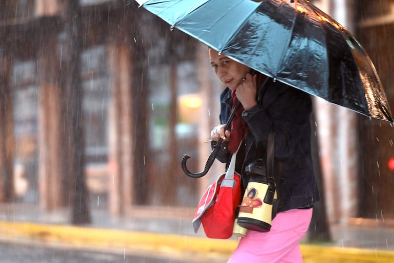 Persona de espaldas con camiseta negra inspecciona vehículo volcado. Camioneta policial con luces encendidas en un día lluvioso.
