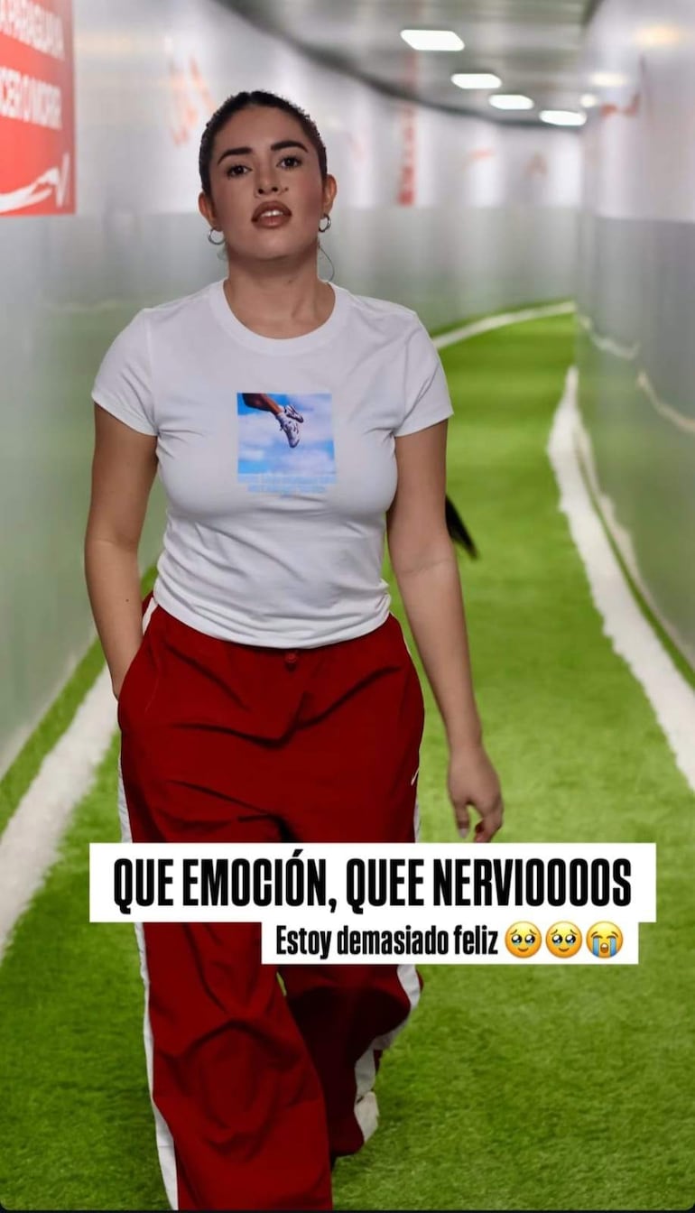 Mujer con camiseta blanca y pantalones rojos sonríe mientras camina por un pasillo, rodeada de decoraciones deportivas.
