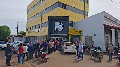 Grupo de personas en fila frente a un edificio amarillo, conversando entre sí bajo un día soleado, con un automóvil blanco estacionado.