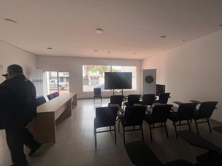Hombre con abrigo y gafas oscuras se aleja de la cámara en sala con banners de IUSS y mesa de conferencias.