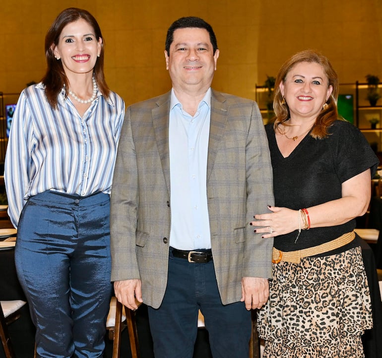 Dos mujeres y un hombre sonríen en un evento, una mujer con blusa de rayas y otra con blusa negra y falda estampada.