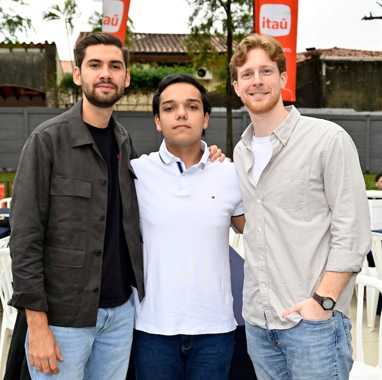 Tres hombres sonríen en celebración, uno en chaqueta negra, otro en camiseta blanca, y el último en camisa gris claro, rodeados de mesas y banderas.