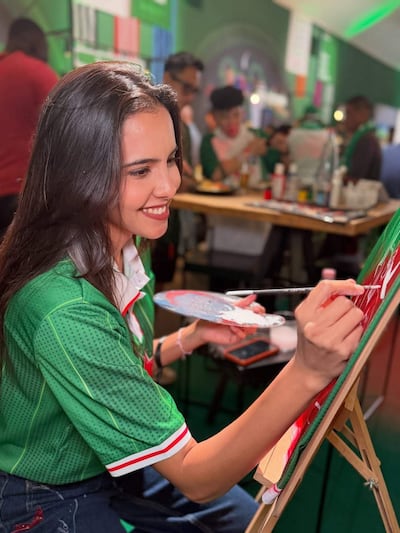 Mujer con camiseta verde y detalles rojos y blancos, sonríe mientras pinta, rodeada de personas festivas en el fondo.