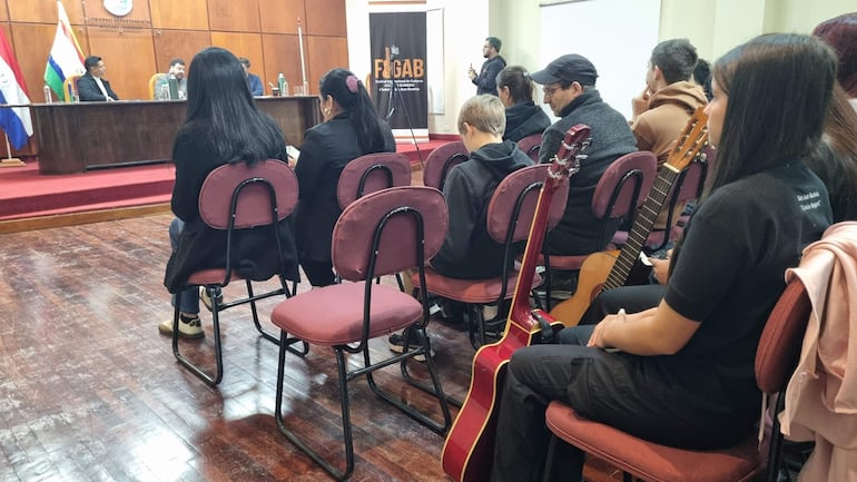 Artistas jóvenes sentados en una mesa con banderas, escuchan a un presentador mientras sostienen guitarras y otros instrumentos.