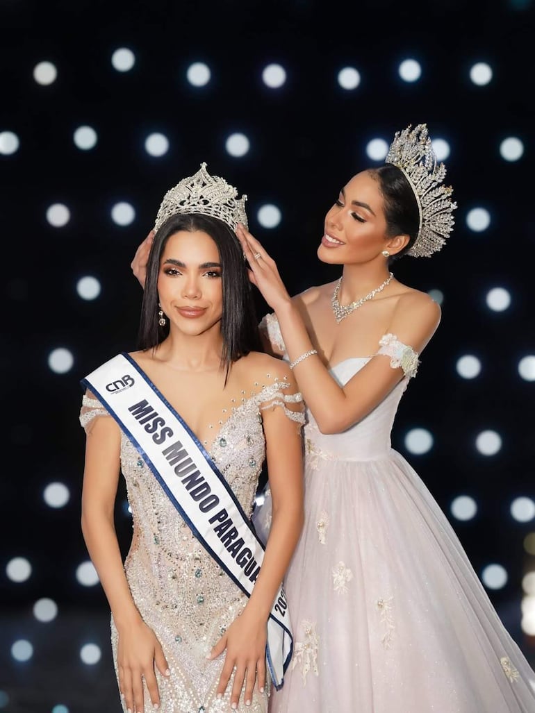 Mujer con banda de Miss Mundo Paraguay 2023 sonríe mientras otra mujer coloca una corona sobre su cabeza en un entorno festivo.