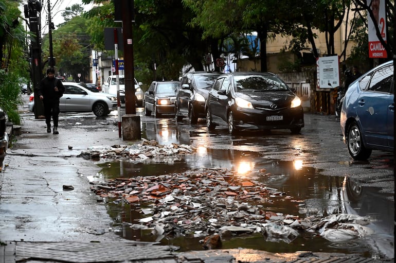 Hombre vestido de negro camina apresurado por la acera, rodeado de tráfico y un charco grande junto a escombros.