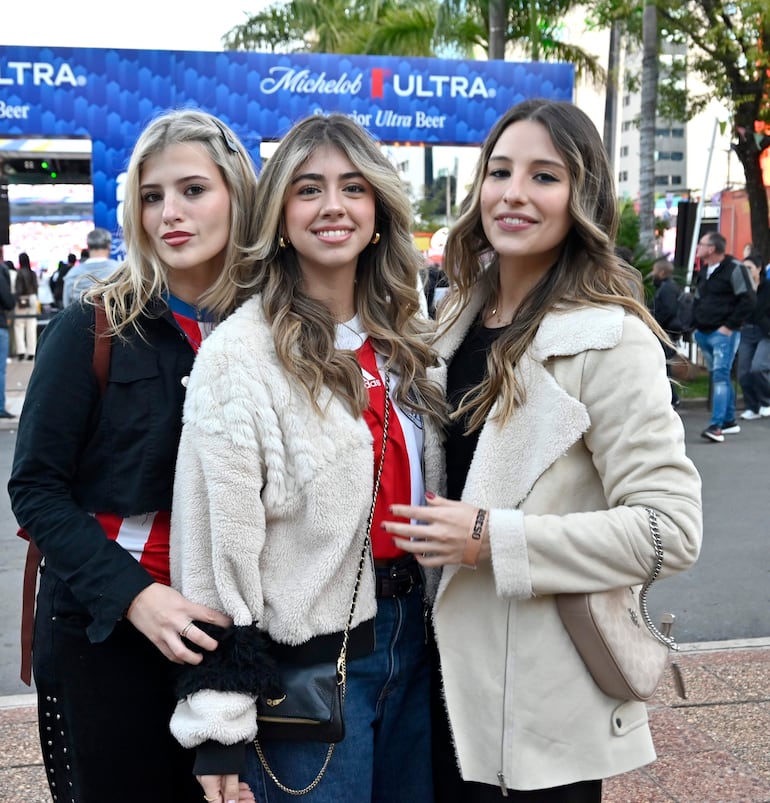 Tres mujeres sonrientes, una en camiseta roja, posan juntas en evento festivo, con cartel de Michelob Ultra al fondo.