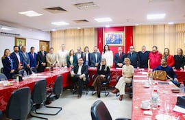Grupo de personas de pie y sentadas, principalmente en vestimenta formal, en una sala de reuniones con una mesa y un retrato político detrás.