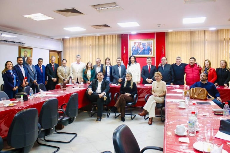 Grupo de personas de pie y sentadas, principalmente en vestimenta formal, en una sala de reuniones con una mesa y un retrato político detrás.