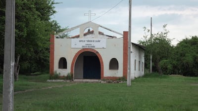 Capilla 'Virgen de Luján' con paredes blancas y cruz en la parte superior, rodeada de césped y árboles en un entorno tranquilo.